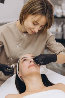 Cosmetician wearing black gloves applying a facial mask to a young woman lying on a stretcher in a beauty salon
