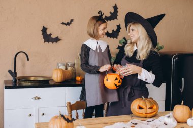 Mother wearing witch costume putting candies into a pumpkin bucket held by her daughter, in their kitchen decorated for Halloween