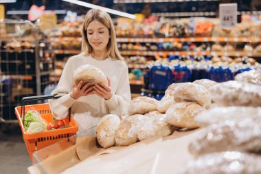 Müşteri süpermarket vitrininden bir somun ekmek seçiyor, elinde taze ürünlerle bir alışveriş sepeti tutuyor.