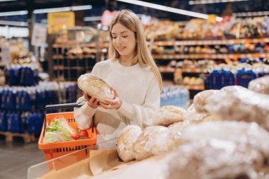 Bir marketin fırın bölümünden ekmek seçen genç bir kadın, elinde taze ürünlerle bir alışveriş sepeti tutuyor.