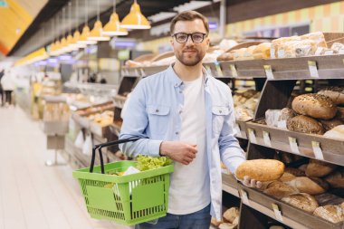 Customer buying bread in supermarket bakery section holding a shopping basket with fresh vegetables