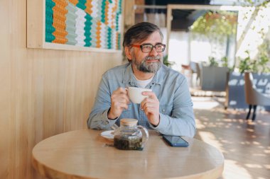 Bearded senior man enjoying a cup of tea in a modern cafe, taking a break and looking away thoughtfully