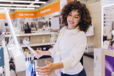 Smiling curly haired woman choosing electric toothbrush in appliance store, comparing different models and brands