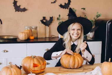 Young woman wearing a witch hat joyfully carving pumpkins for a Halloween party in a cozy kitchen, embracing the festive autumn spirit