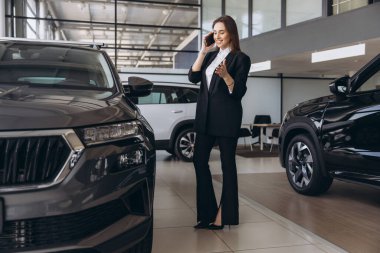Saleswoman in a tailored suit engaging in conversation on the phone while showcasing a new car to an interested customer in a dealership showroom