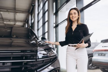 Smiling saleswoman holding clipboard presenting new car to customer in car dealership
