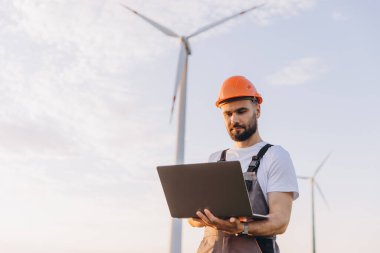 Engineer wearing a hardhat and using a laptop, working diligently on a wind turbine to enhance renewable energy production and sustainability