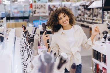 Smiling woman with curly hair testing a new hair straightener while shopping in an appliance store, enjoying the selection of beauty products