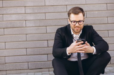 Businessman looking at smartphone screen with worried expression, sitting on sidewalk against brick wall