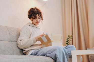 Happy customer joyfully opening a cardboard box, revealing a newly delivered book while relaxing on her sofa in the cozy living room