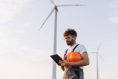 Arab male engineer holding an orange helmet and using a digital tablet while working on a wind turbine farm for sustainable energy production