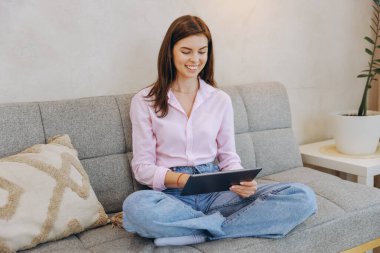 Smiling woman using digital tablet while sitting comfortably on sofa at home, enjoying leisure time and online activity