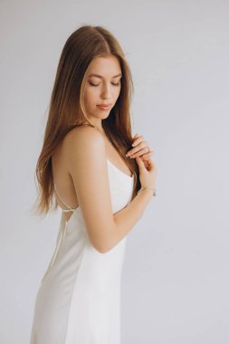 Studio portrait of a young woman with long brown hair wearing a white dress, gently touching her hair