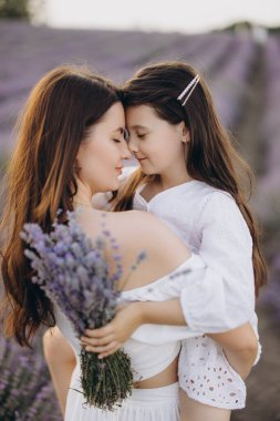 Mother holding her daughter in her arms, both wearing white dresses, touching foreheads with closed eyes in a lavender field, holding a bouquet of lavender