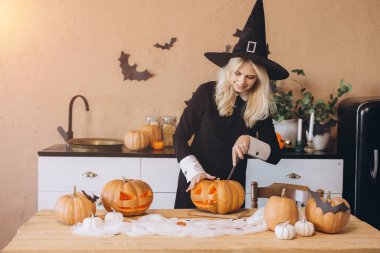Young woman wearing a witch hat, joyfully carving pumpkins for a Halloween party in a festively decorated kitchen, embracing autumn vibes