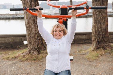 Elderly woman working out on a pull up machine at an outdoor gym, promoting healthy aging and active lifestyle