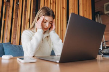 Young woman experiencing fatigue and headache while working remotely on a laptop in a cozy cafe, surrounded by natural light and quiet