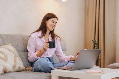 Happy freelancer enjoying coffee while working on a laptop from a cozy sofa, balancing productivity and relaxation in a home office setting