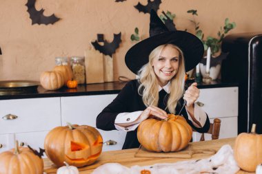Smiling woman wearing a witch hat joyfully carving a pumpkin for a festive Halloween party celebration in a cozy kitchen