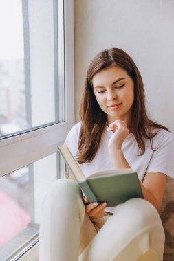 Serene young woman relaxing by the window, engrossed in a captivating book, finding peace and tranquility in literature