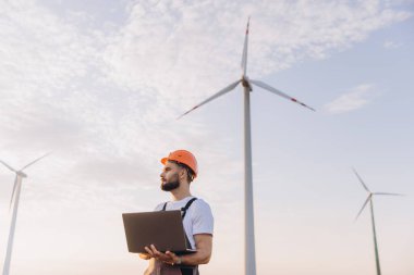Engineer with safety helmet holding laptop inspecting wind turbines in a wind farm for renewable and sustainable energy production