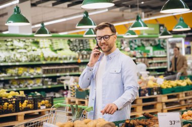 Man selecting fresh potatoes while engaged in a phone conversation and pushing a shopping cart through the supermarket aisles