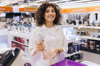 Smiling curly haired woman holding handheld garment steamer reading instruction manual in home appliance store