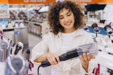 Smiling woman with curly hair selecting a new hair dryer while shopping in an electronics store filled with various appliances