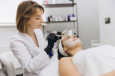 Professional beautician applying a clay mask treatment to a young woman's face in a beauty salon