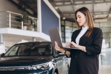 Saleswoman standing next to a car in a dealership, using a laptop to assist customers with online browsing and vehicle information