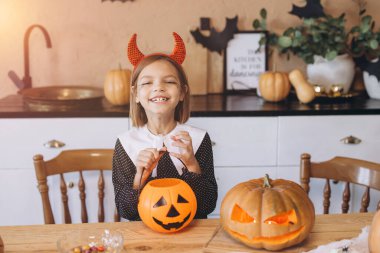 Little girl wearing devil horns headband smiling and holding a pumpkin basket in a kitchen decorated for Halloween