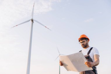 Arabian engineer wearing an orange helmet and glasses reading blueprints on a wind turbine farm, concept of renewable energy