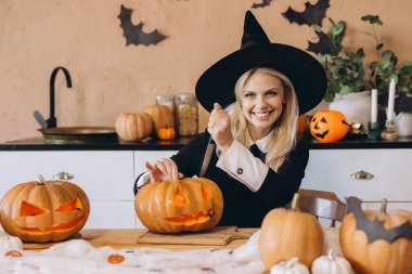 Young woman wearing witch hat carving a pumpkin for Halloween, with other decorated pumpkins around her