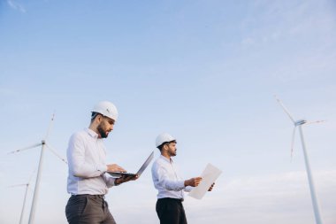 Two engineers working on a wind turbine project using laptop and blueprints under a clear blue sky
