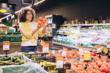 Young curly woman choosing tomatoes in a supermarket, holding a bunch of tomatoes in her hands and smiling