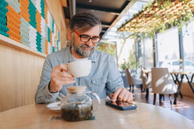 Bearded senior man sipping tea while using a smartphone, enjoying a peaceful moment in a modern cafe filled with natural light