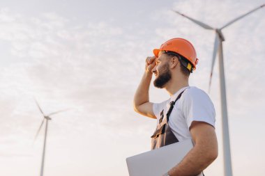 Arab engineer holding a laptop while inspecting wind turbines in the desert, focusing on sustainable energy solutions for the future