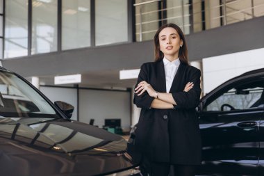 Saleswoman standing confidently with arms crossed in front of a sleek car, showcasing professionalism in a modern car dealership showroom