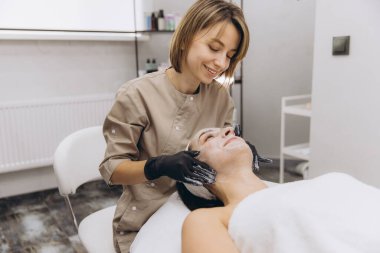 Smiling beautician wearing black gloves applying a facial mask to a client lying on a bed in a beauty salon