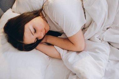 Young woman resting peacefully in bed, surrounded by soft white bedding, enjoying a serene and comfortable sleep in a cozy bedroom
