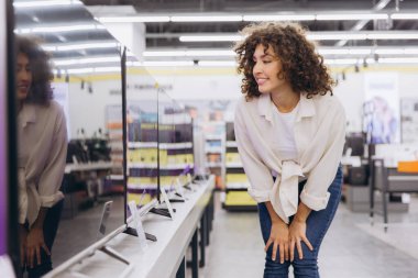 Smiling woman with curly hair selecting a new television in a modern consumer electronics store, excited about the variety of options available