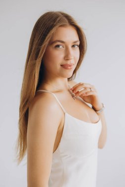 Studio portrait of a young woman with long brown hair, wearing a white dress and necklace, touching her chest