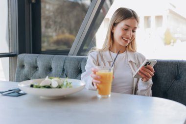 Young blonde woman using mobile phone while having a healthy lunch with salad and orange juice in a modern restaurant