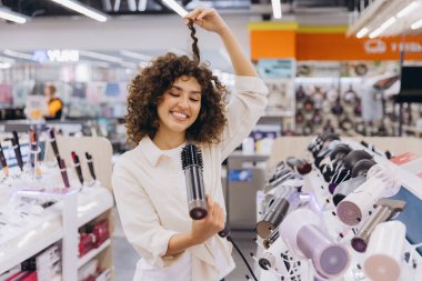 Smiling woman with curly hair testing new hair dryer in appliance store, enjoying shopping experience