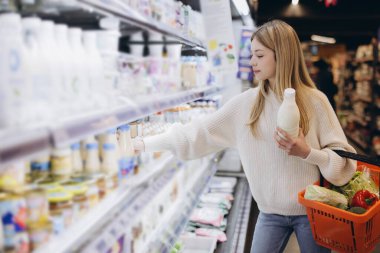 Müşteri dondurulmuş raflardan yoğurt seçiyor, elinde süt şişesi ve market alışverişi yapıyor, süpermarkette haftalık alışverişini yapıyor.