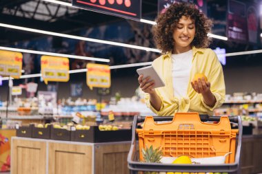 Smiling woman checking shopping list on notepad and choosing oranges while buying groceries in supermarket