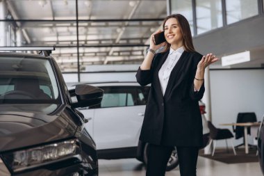 Smiling saleswoman talking on the phone while presenting a new car inside a modern car dealership