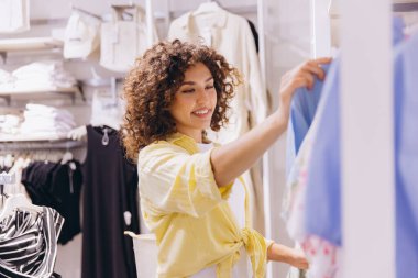 Smiling woman with curly hair enjoying a shopping experience while selecting stylish clothes in a vibrant fashion retail store