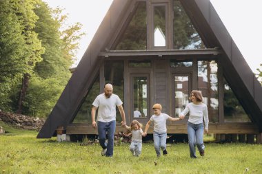 Happy family running on the grass in front of their modern, sustainable a frame cabin, enjoying a weekend in the woods