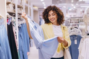 Smiling woman with curly hair joyfully holding two blue shirts while shopping in a vibrant fashion store, enjoying the retail experience
